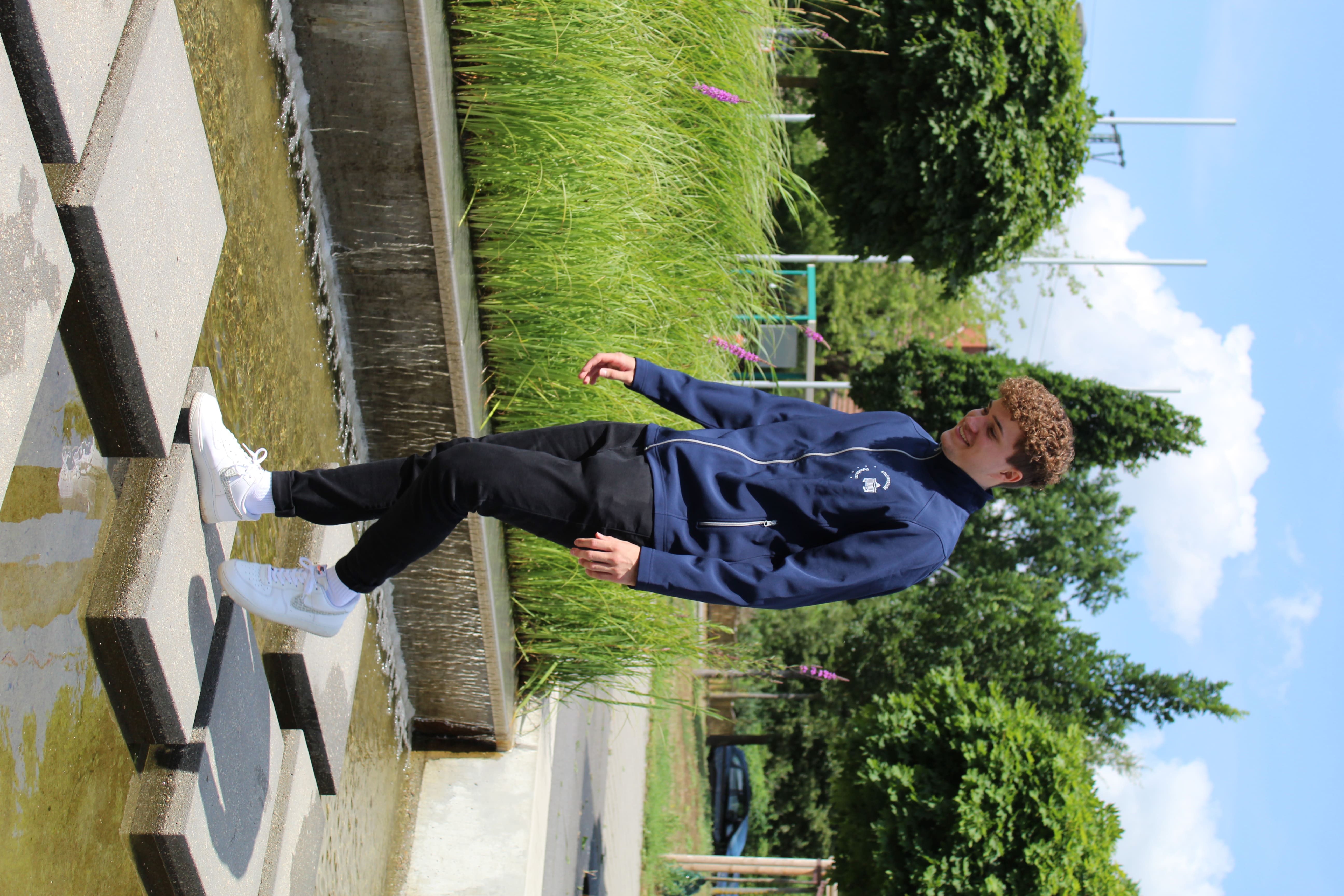 Softshelljacke-Herren. Abgebildet ist ein junger Mann mit einer blauen Softshelljacke. Er läuft auf Steinplattformen zwischen denen Wasser fließt.
