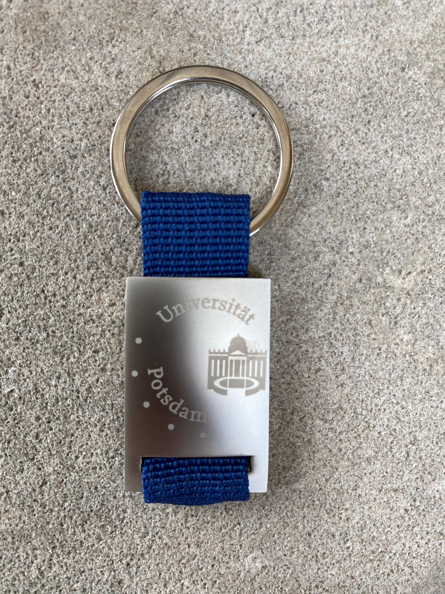 Schlüsselanhänger-Logo. Abgebildet ist ein Schlüsselanhänger aus Edelstahl mit einem blauen Band und dem Universitätslogo.
