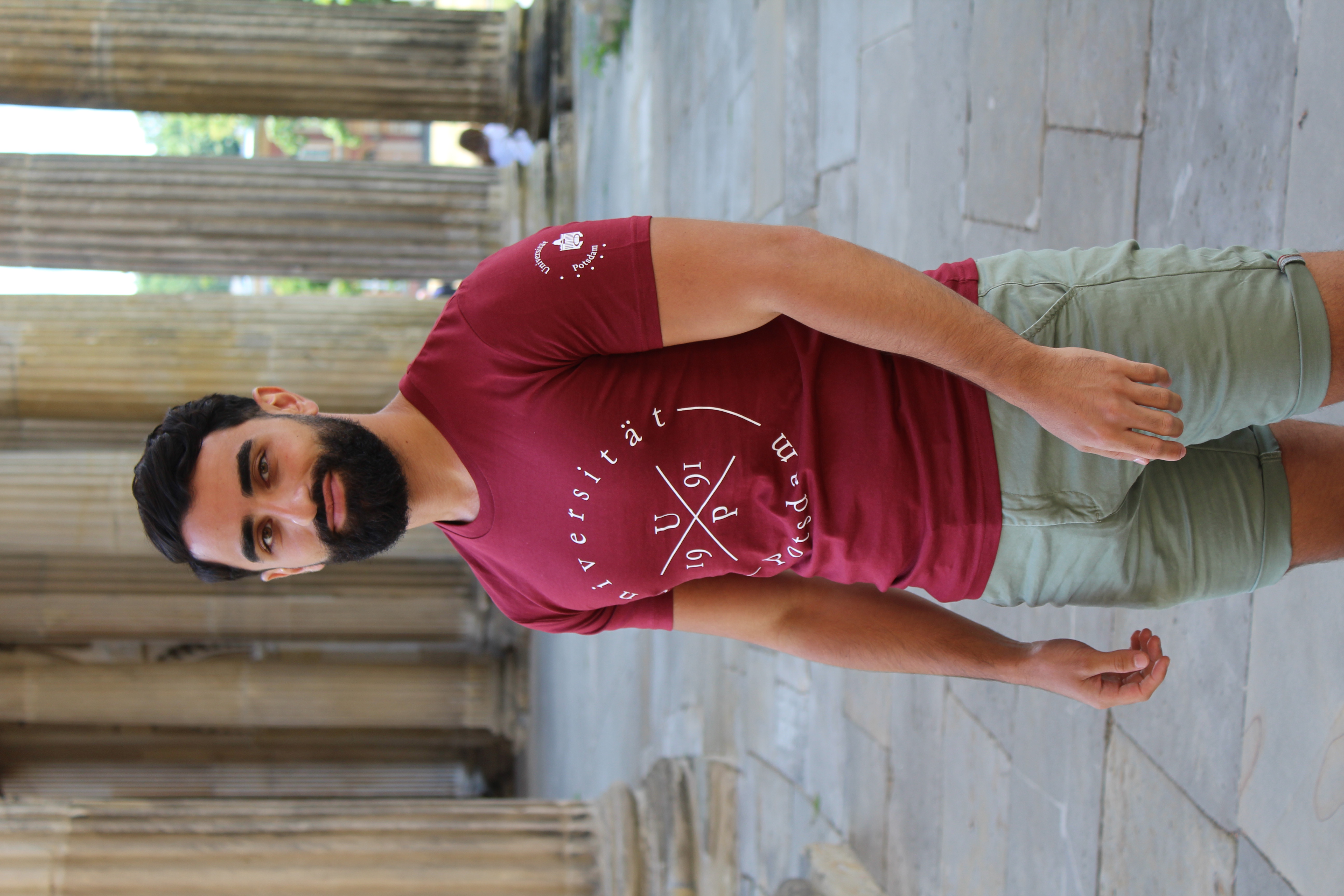 Styler T-shirt. Abgebildet ist ein junger Mann, der im Freien vor Säulen posiert. Er trägt ein burgunder farbenes T-shirt im Design Styler.