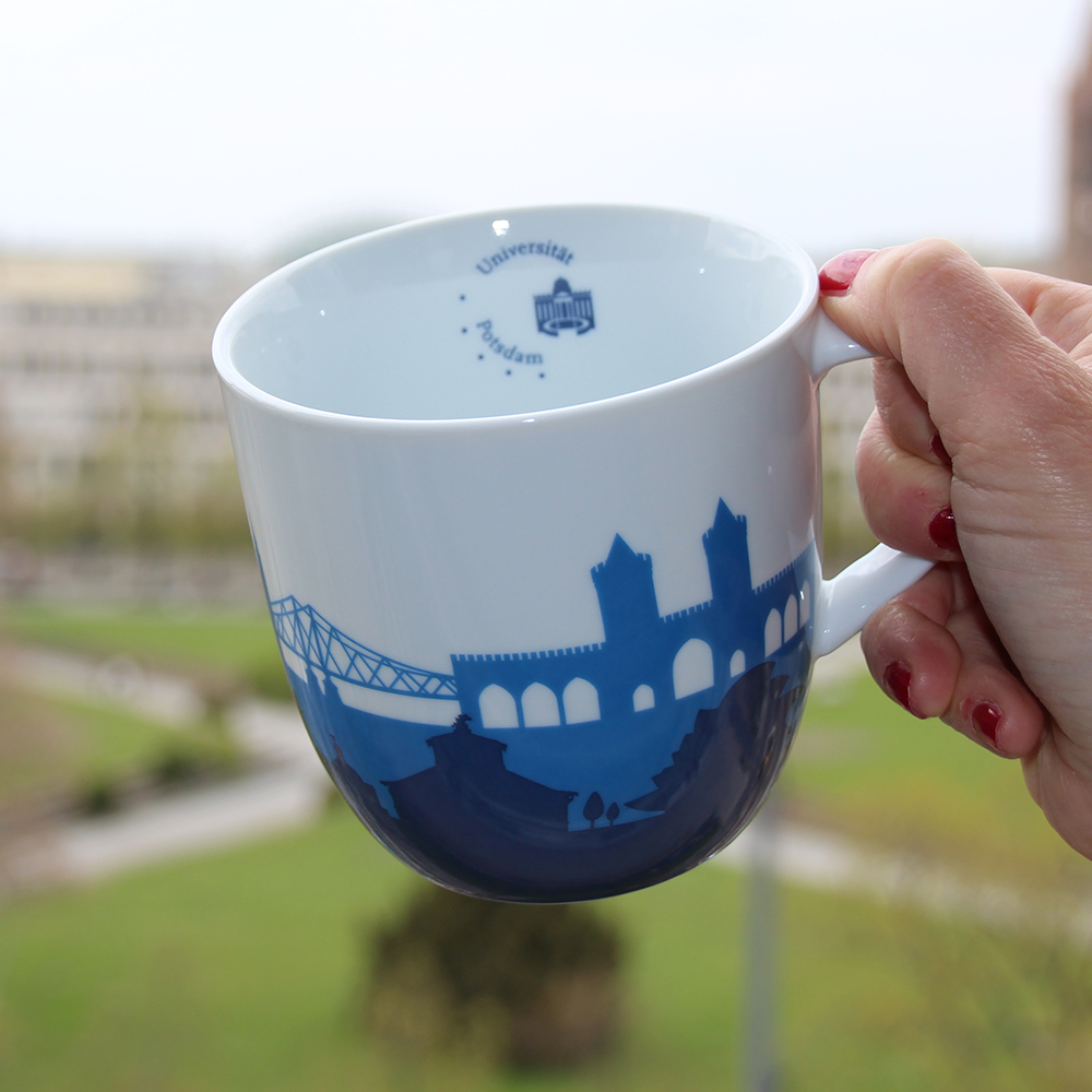 Jumbo-tasse. Abgebildet ist eine große weiße Tasse mit einem blauen Brückenlogo. Im Hintergrund sieht man Wiesen und Wege.